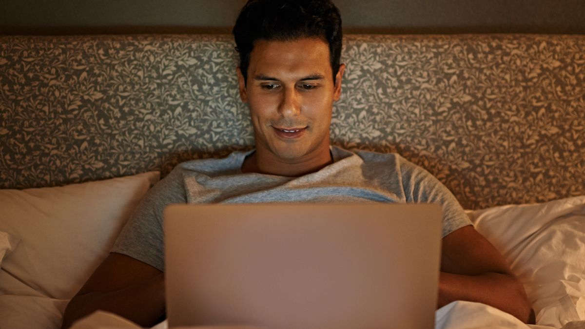Egy férfi az ágyban ülve laptopot használ, mosolyogva néz a képernyőre.