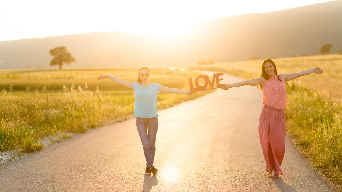 Két nő boldogan sétál egy naplementés úton, kezükben a 'LOVE' feliratú táblával.